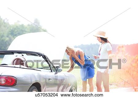Picture - Female friends examining broken down car on country road. Fotosearch