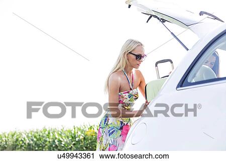 Stock Image - Female friends loading luggage in car trunk against clear sky. Fotosearch