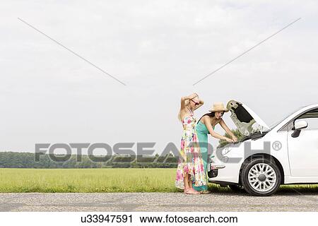 Stock Image - Friends examining broken down car on country road against clear sky. Fotosearch