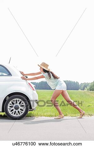 Stock Image - Full-length of woman pushing broken down car on country road. Fotosearch