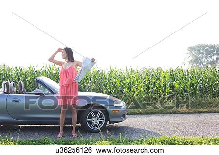 Stock Photograph - Full length of woman shielding eyes while holding map by convertible. Fotosearch