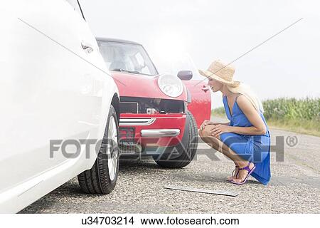 Picture - Full-length side view of tensed woman looking at damaged cars on road. Fotosearch