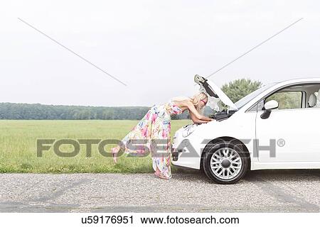 Stock Image - Full-length side view of woman examining broken down car on country road. Fotosearch