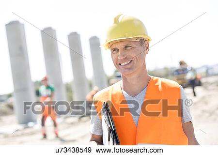 Happy architect looking away while holding clipboard at construction site View Large Photo Image Stock Photo - Happy architect looking away while holding clipboard at construction site. Fotosearch