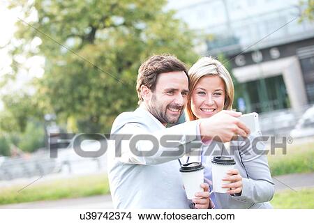 Happy business couple taking selfie while holding disposable cups in city View Large Photo Image Picture - Happy business couple taking selfie while holding disposable cups in city. Fotosearch