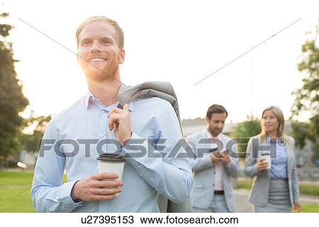 Stock Image - Happy businessman holding disposable cup with colleagues standing in background. Fotosearch