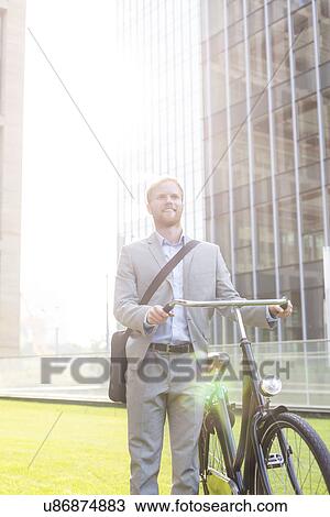 Stock Image - Happy businessman standing with bicycle outside building. Fotosearch