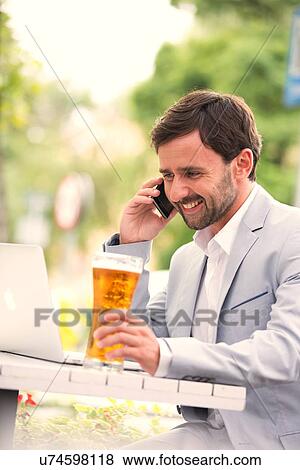 Stock Photo - Happy businessman using mobile phone and laptop while holding beer glass at outdoor restaurant. Fotosearch