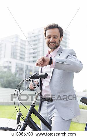 Stock Image - Happy businessman with bicycle checking time outdoors. Fotosearch