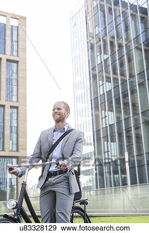 Stock Photo - Happy businessman with bicycle standing outside office building. Fotosearch