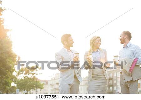 Stock Image - Happy businesspeople with disposable cups talking against clear sky on sunny day. Fotosearch