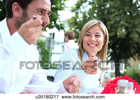 Stock Photo - Happy businesswoman eating food with colleague at sidewalk cafe. Fotosearch