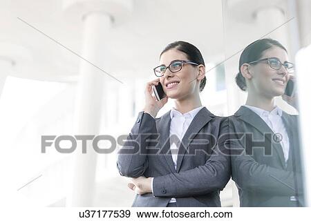 Happy businesswoman using cell phone while leaning on glass wall View Large Photo Image Stock Photo - Happy businesswoman using cell phone while leaning on glass wall. Fotosearch