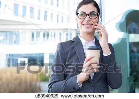 Stock Photo - Happy businesswoman using cell phone while holding disposable cup outdoors. Fotosearch