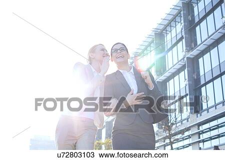 Happy businesswoman whispering in colleague's ear outside office building on sunny day View Large Photo Image Stock Image - Happy businesswoman whispering in colleague's ear outside office building on sunny day. Fotosearch