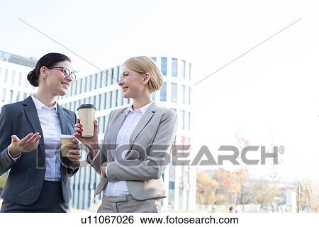 Stock Photograph - Happy businesswomen conversing while holding disposable cups outside office building. Fotosearch