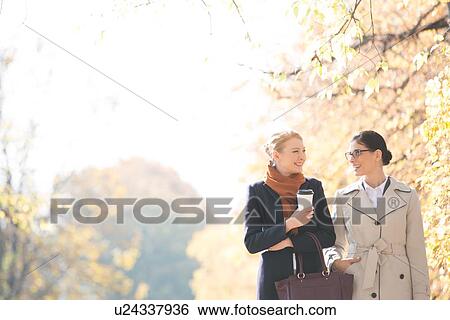 Stock Photograph - Happy businesswomen conversing while walking at park on sunny day. Fotosearch