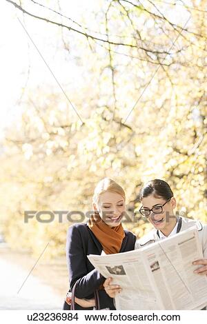 Picture - Happy businesswomen reading newspaper together at park. Fotosearch