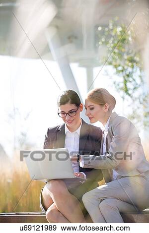 Stock Photo - Happy businesswomen using laptop together on sunny day. Fotosearch
