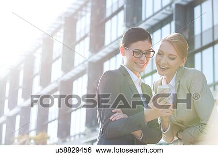 Stock Photograph - Happy businesswomen using smart phone outside office building on sunny day. Fotosearch