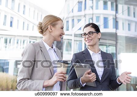 Happy businesswomen with tablet PC and disposable cup conversing outside office building View Large Photo Image Stock Photo - Happy businesswomen with tablet PC and disposable cup conversing outside office building. Fotosearch