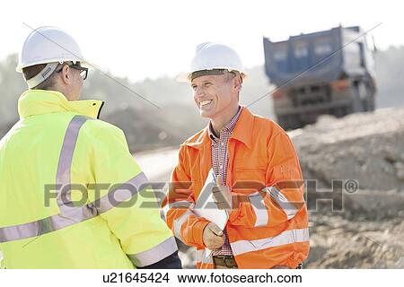 Picture - Happy engineer discussing with colleague at construction site on sunny day. Fotosearch
