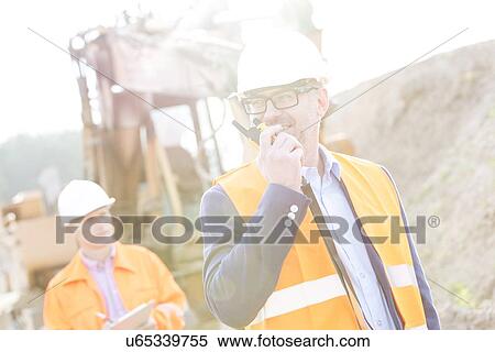 Stock Photography - Happy engineer using walkie-talkie on construction site with colleague in background. Fotosearch