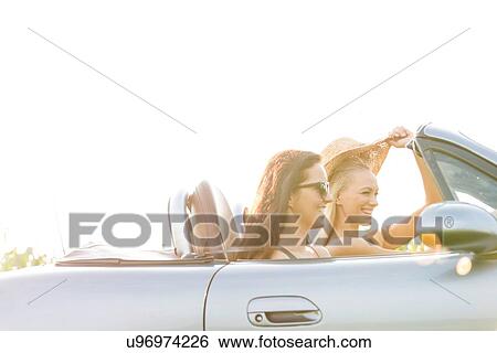 Stock Photograph - Happy female friends enjoying road trip in convertible against clear sky. Fotosearch