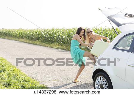 Happy female friends pushing luggage in car trunk against clear sky View Large Photo Image Stock Photo - Happy female friends pushing luggage in car trunk against clear sky. Fotosearch