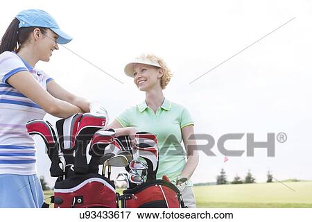 Stock Photo - Happy female golfers talking at golf course against clear sky. Fotosearch