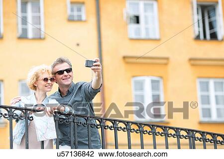 Stock Image - Happy middle-aged couple taking selfie through camera against building. Fotosearch
