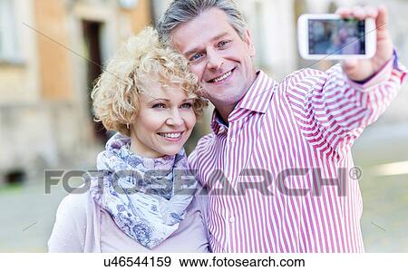 Happy middle-aged couple taking selfie through smart phone outdoors View Large Photo Image Stock Photo - Happy middle-aged couple taking selfie through smart phone outdoors. Fotosearch