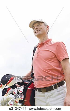 Stock Image - Happy middle-aged man looking away while carrying golf bag against clear sky. Fotosearch