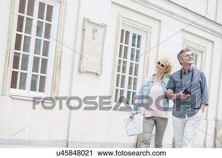 Stock Image - Happy middle-aged tourist couple walking arm in arm by building. Fotosearch