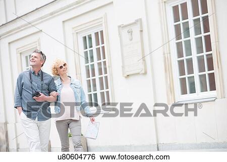 Happy middle-aged tourist couple walking arm in arm by building View Large Photo Image Stock Photograph - Happy middle-aged tourist couple walking arm in arm by building. Fotosearch