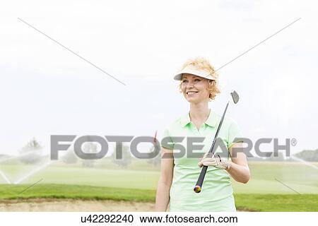 Stock Image - Happy middle-aged woman looking away while holding golf club. Fotosearch