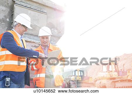 Stock Image - Happy supervisors discussing over clipboard at construction site against clear sky. Fotosearch