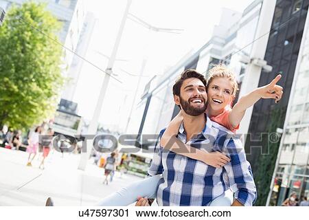 Stock Image - Happy woman showing something to man while enjoying piggyback ride in city. Fotosearch
