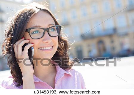 Stock Image - Happy young woman looking away while using smart phone on sunny day. Fotosearch