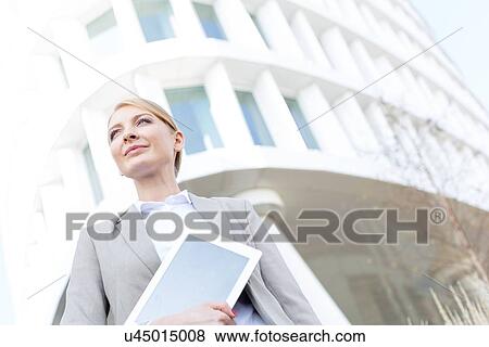 Stock Photo - Low angle view of confident businesswoman holding digital tablet outside office building. Fotosearch