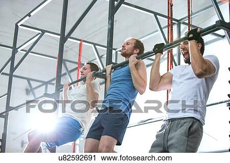 Stock Image - Low angle view of dedicated men doing chin-ups in crossfit gym. Fotosearch