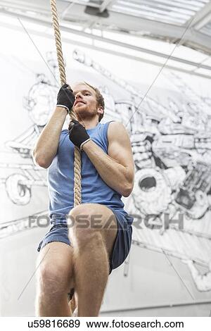 Stock Photo - Low angle view of determined man climbing rope in crossfit gym. Fotosearch