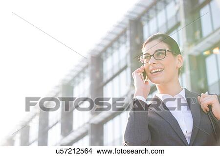 Picture - Low angle view of happy businesswoman using cell phone outside office building. Fotosearch