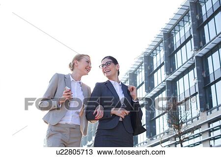 Stock Image - Low angle view of happy businesswomen walking outside office building against clear sky. Fotosearch