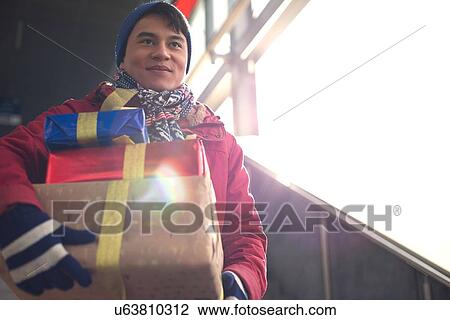 Stock Image - Low angle view of smiling man holding gifts by window. Fotosearch