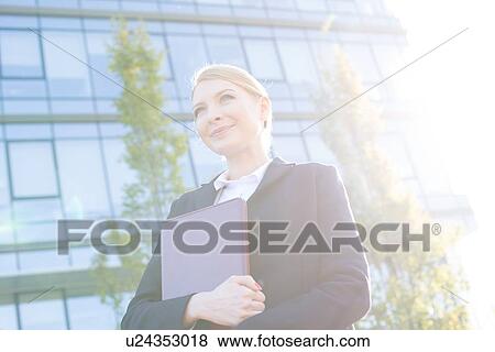 Stock Photo - Low angle view of thoughtful businesswoman holding folder on sunny day. Fotosearch