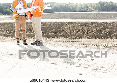 Stock Image - Low section of engineers with blueprints standing at construction site. Fotosearch