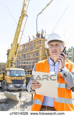 Stock Photo - Male supervisor using walkie-talkie while holding clipboard at construction site. Fotosearch