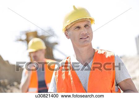 Stock Image - Male worker at construction site with colleague standing in background. Fotosearch