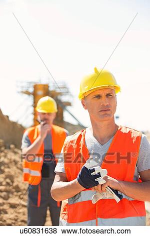 Stock Photo - Male worker standing at construction site with colleague in background. Fotosearch